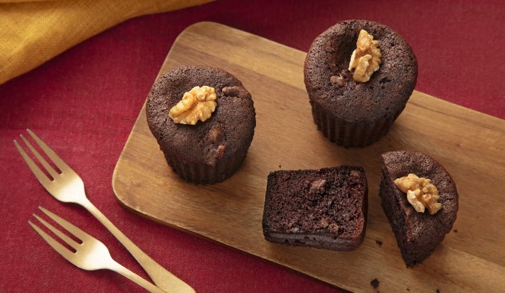 Fotografia vista de cima de três muffins de nescau sobre uma tábua de madeira retangular, ao lado de dois garfos dourados. Um dos muffins está cortado na metade para mostrar a maciez da massa.