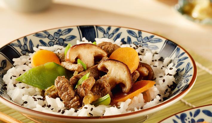 fotografia em tons de branco de bege de uma bancada bege vista de frente. Contém dois recipientes redondos em tons de branco e azul com arroz, cenouras fatiadas e shitake, ao lado o hashi para servir e ao fundo uma xicara branca.