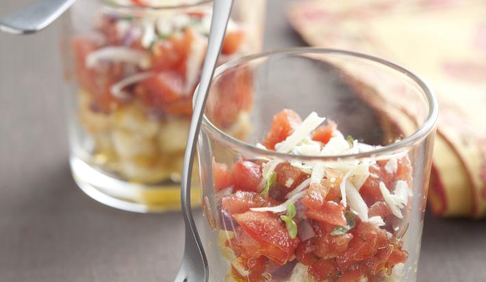 Fotografia em tons de amarelo e vermelho em uma bancada cinza com uma toalha com detalhes amarelos e vermelho. Ao centro, dois copinhos de vidro com a bruschetta com tomate, croutons e queijo dentro deles.