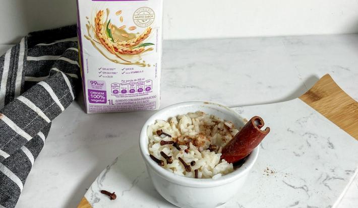 Foto em tons claros da receita de arroz doce cremoso servida em um bowl branco de porcelana sobre uma tábua de mármore branco. Ao fundo, há uma caixa de bebida vegetal de aveia natures heart e um paninho preto