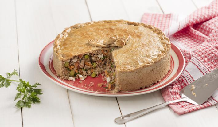 Fotografia em tons de branco, dourado e vermelho, com torta de carne assada e fatiada sobre prato vermelho e branco, ao lado guardanapo em branco e vermelho e uma espátula, ramo de salsa ao lado esquerdo, tudo sobre bancada de madeira na cor branca.