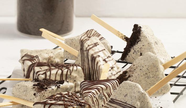 Fotografia em tons de branco em uma bancada de cor branca. No centro, estão os Cakes Pop Cookies’n Cream polvilhados com chocolate. Ao fundo, um recipiente contendo Topping de Negresco.
