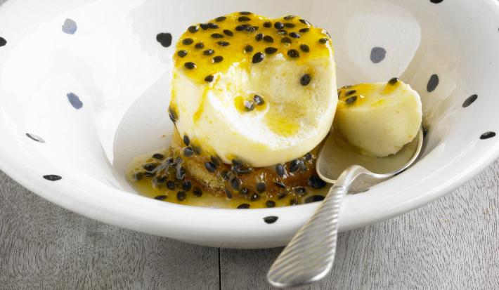 Fotografia de um flan de maracujá e manga, com calda de maracujá em cima e no fundo do prato, que tem cor branca com bolinhas pretas. Apoiado no prato, uma colher de sopa segurando um pedaço do flan. A sobremesa está sobre uma mesa de madeira cinza.