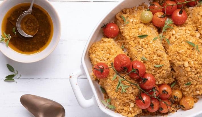 Fotografia em tons de amarelo em uma bancada de madeira branca com um refratário oval branco com as postas de bacalhau com crosta de pão e tomatinhos cereja assados dentro dele.