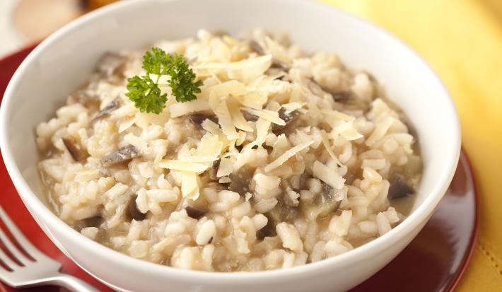 Fotografia em tons de cinza, branco, vermelho e amarelo de uma bancada de madeira cinza vista de cima. Um prato vermelho contém um recipiente redondo branco por cima com risoto, ao lado um garfo e do outro lado um pano amarelo e ao fundo uma taça.