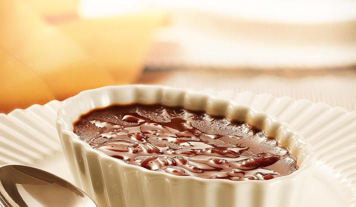 fotografia em tons de branco e marrom de uma bancada vista de frente, contém um prato redondo e branco com um potinho oval branco com um creme de chocolate dentro e uma colher ao lado para servir.