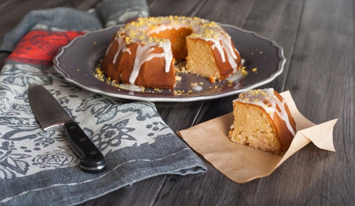 Fotografia de um bolo de bundt parcialmente fatiado, exibindo sua textura densa e úmida. Coberto com um glacê brilhante e raspas de limão, está disposto em um prato escuro, ao lado de uma faca e sobre um pano decorativo.
