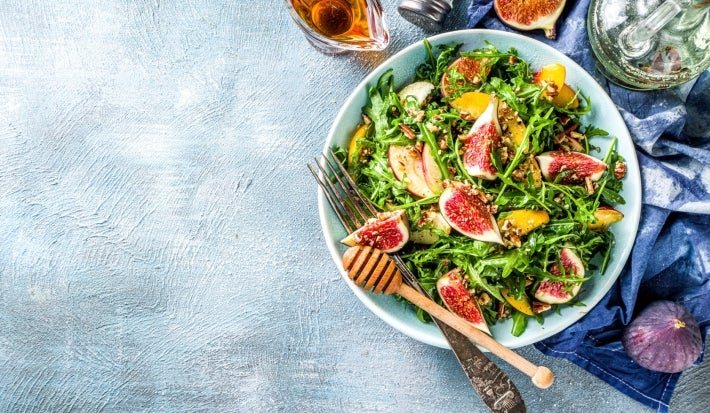 Em cima de uma mesa, prato com salada com mix de folhas, frutas em cubos e figo.