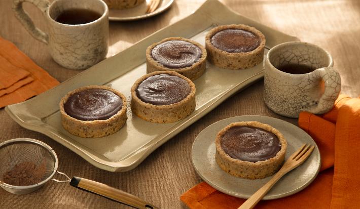 Foto em tons de marrom da receita de torta de ganache de cacau servida em diversas porções sobre uma base de cerâmica retangular e uma sobre um pires de cerâmica, além de duas caneca de mesmo material ao lado