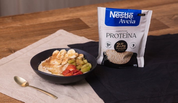 Receita de mingau com banana, morango, kiwi e mel, acompanhado de colher dourada e uma embalagem de nestlé aveia proteína