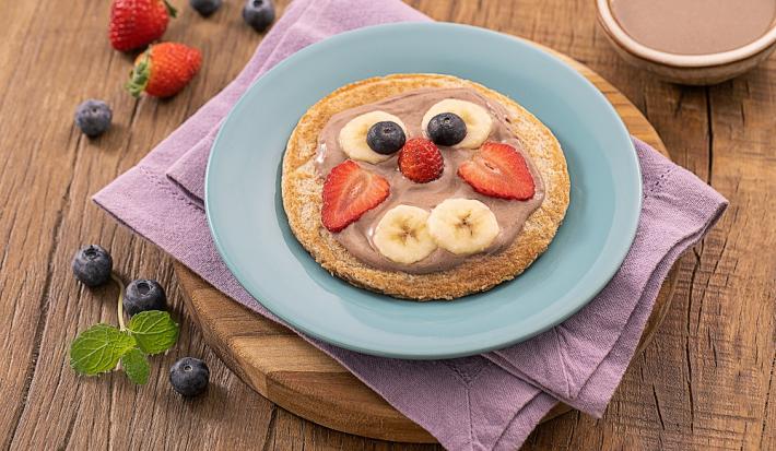 Foto da receita de panquequinha de cenoura em formato de "rostinho" com as frutas, servida em um prato de porcelana azul claro sobre um paninho lilás em uma tábua de madeira. Ao fundo algumas frutas espalhadas e um bowl de porcelana com cacau em pó.