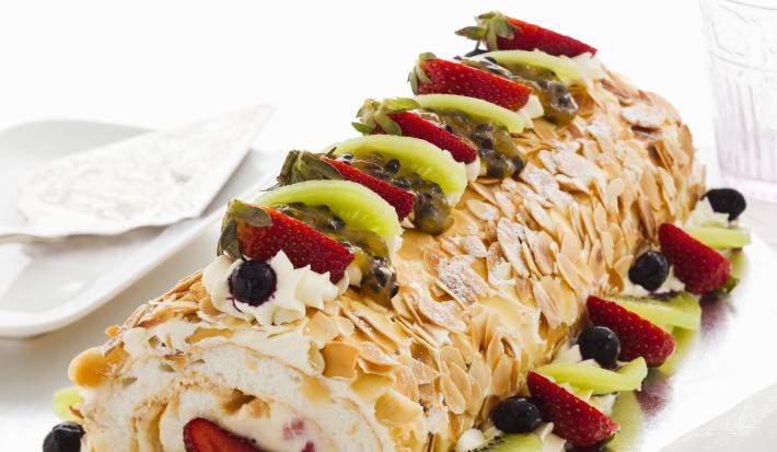 Fotografia de um rocambole com geleia, recheado com sorvete, coberto por lâminas de amêndoas, merengue e frutas, em cima de um apoio prateado, sobre uma mesa branca. Ao lado, um copo e um prato pequeno de sobremesa, com um guardanapo por cima.