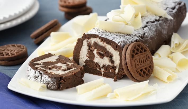 Fotografia em tons de azul em uma bancada de madeira azul, um pano azul escuro, um prato quadrado branco raso com o rocambole de chocolate com recheio de Leite Moça e raspas de chocolate branco em cima do prato. Ao lado, Biscoitos Negresco espalhados.