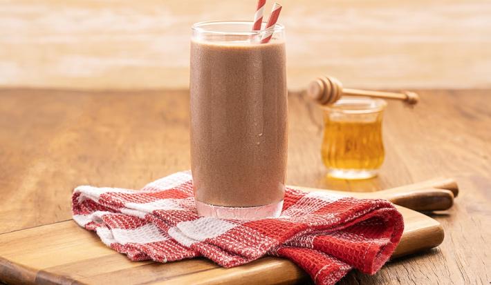Foto da receita de milkshake proteico com abobrinha e cacau servida em um copo alto e largo com dois canudos listrados, embaixo um pano quadriculado vermelho e branco sobre uma tábua de madeira. Ao fundo um copo do vidro com mel