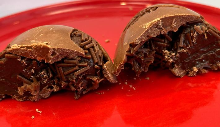 Na imagem duas cascas de ovo de pascoa de chocolate recheadas com brigadeiro em tons marrom e castanha. As cascas estão sobre um prato vermelho que preenche todo o restante da imagem.