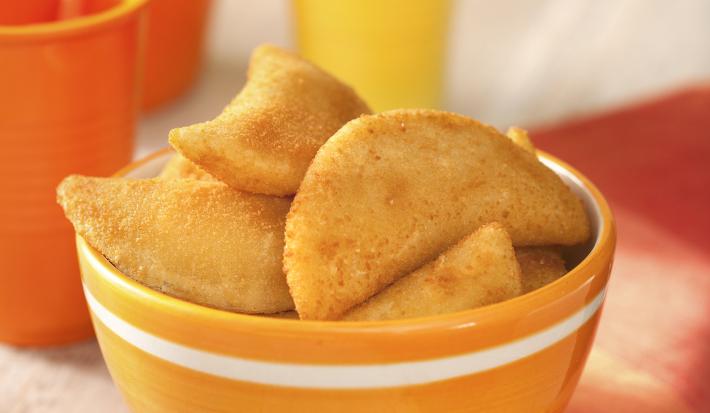 Fotografia em tons de amarelo e laranja, ao centro potinho com rissoles fritos, ao lado guardanapo laranja, acima copo amarelo com canudo, no lado esquerdo jarra e copo laranja com um canudo, tudo sobre bancada de madeira em pátina.