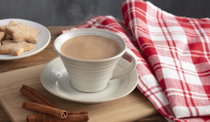 Fotografia em tons de vermelho e marrom de uma bancada, ao centro uma xícara com chocolate quente saindo fumaça e um pires abaixo. Ao lado pedaços de canela, biscoitinhos e uma pano xadrez vermelho e branco.