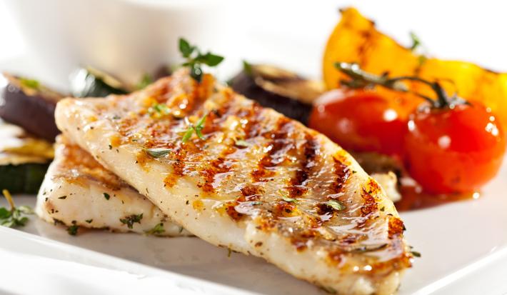 Fotografia de duas postas de peixe no azeite com alguns legumes no fundo, sobre um prato branco grande.