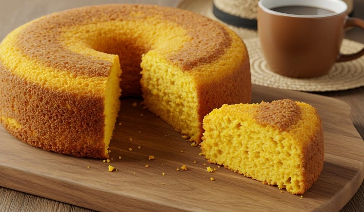 Fotografia de um bolo de milho recém-assado, com textura macia e esponjosa. Apresenta uma crosta dourada e um furo no centro, servido em uma tábua de madeira, ao lado de uma xícara de café.