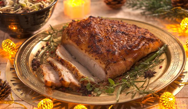 Fotografia em tons de amarelo em uma bancada de madeira, um prato redondo dourado com o pernil em cima dele, ao lado, um potinho com o salpicão. Ao fundo, decoração e enfeites natalinos.