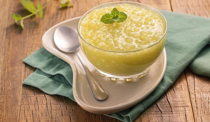 Foto da receita de Sagu Suco Verde. Observa-se uma taça transparente contendo o sagu sobre um prato e um guardanapo verde . Ao lado, uma colherinha e um ramo de hortelã.