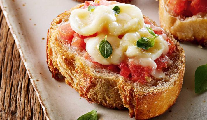 Fotografia em tons de branco e amarelo, com travessa branca e borda dourada com três bruschettas com tomate, mozarela e folhas de manjericão, sobre mesa de madeira rústica.