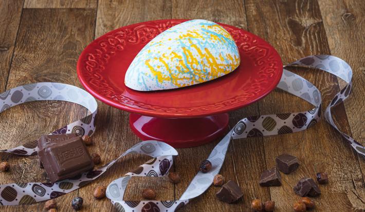 Imagem da receita de uma casca de ovo de páscoa branca, decorada com cores laranja e azul, sobre um prato vermelho, em uma bancada de madeira, decorada com uma fita decorativa, pedaços de chocolate e avelãs