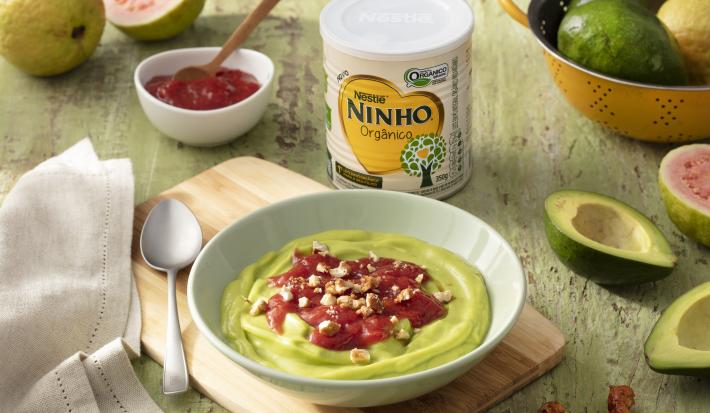 Fotografia em tons de verde de uma bancada verde, sobre ela uma tábua de madeira com um recipiente verde redondo com o creme de abacate e uma colher. Ao lado abacates, um paninho branco, um recipiente com goiabada, goiabas e uma lata de NINHO orgânico.