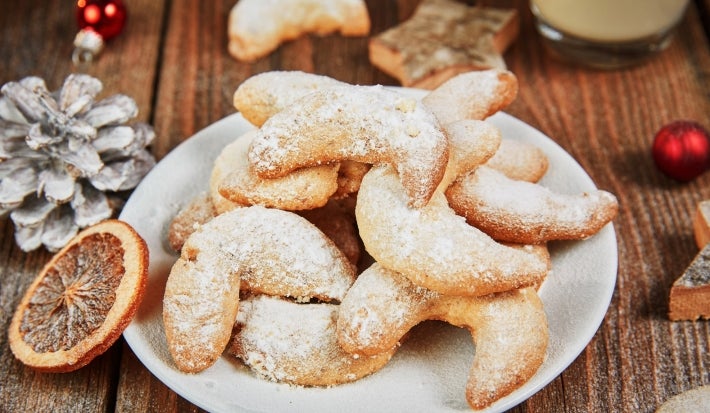 Em cima de uma mesa, prato branco com biscoitos em forma de meia lua, polvilhados com açúcar de confeiteiro.