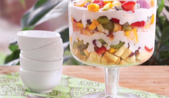em uma bancada contém uma taça de vidro com pedaços de frutas e creme por cima e ao lado uma colher e potinhos brancos empilhados.