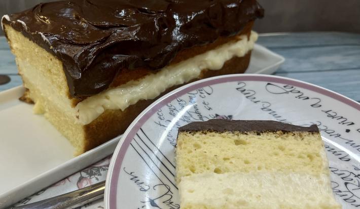 Foto da receita de Boston Pie. Observa-se o bolo retangular com uma camada de creme branco no meio e cobertura de chocolate no plano de fundo. À frente, um prato de sobremesa redondo com uma fatia do bolo.