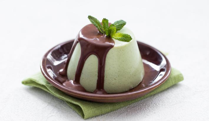 Fotografia de uma mousse de menta com calda de chocolate, com folhas de menta por cima. A mousse está em um prato pequeno na cor marrom, sobre um pano dobrado, na cor verde, que está sobre uma mesa branca.