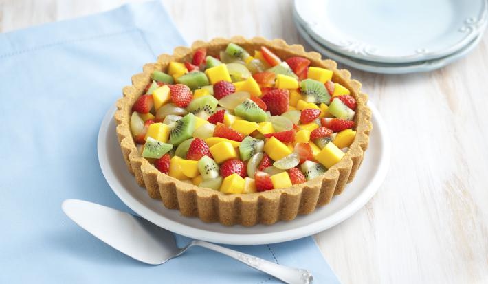 Fotografia em tons de bege, azul, amarelo, vermelho e verde de uma bancada bege vista de cima. Contém um pano azul e por cima um prato redondo branco, com uma torta com frutas picadas (kiwi, manga, morango, uva) por cima.