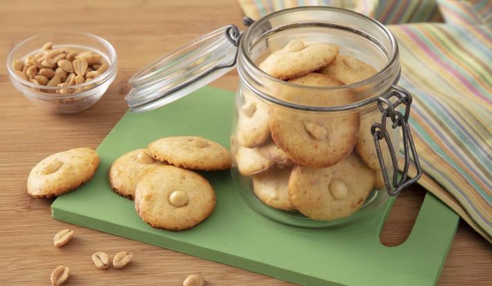 Receita de Crocantinho. Observa-se biscoitos dispostos em uma tábua verde. Ao lado, um pote hermético de vidro com mais biscoitos. Amendoins decoram a foto.
