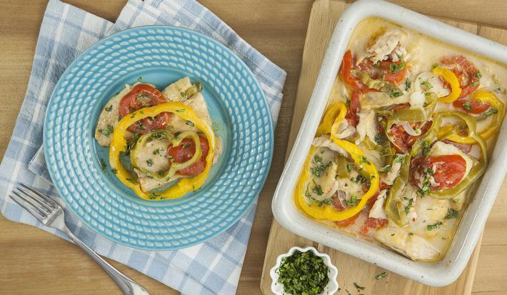 Fotografia em tons de azul em uma bancada de madeira, um pano azul xadrez, um prato azul com um pouco da moqueca de peixe com pimentões vermelho e amarelo, uma tábua de madeira e em cima dela uma travessa com a moqueca com os pimentões.