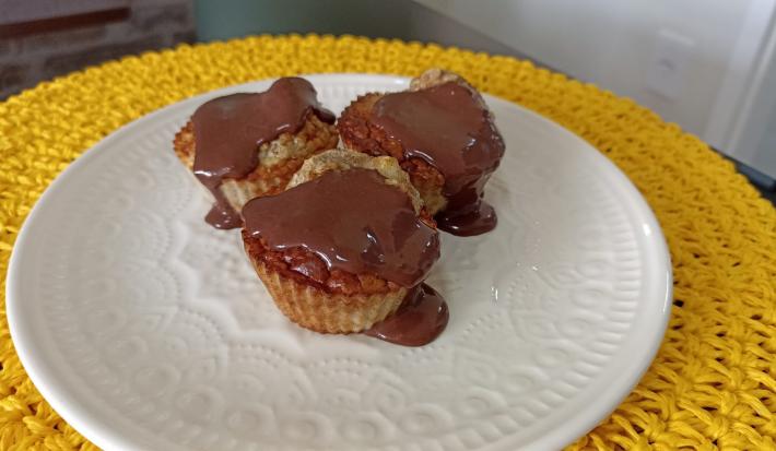 Fotografia em tons de amarelo em uma bancada de pedra, um apoia prato amarelo de renda, um prato branco com três bolinhos de banana com calda de chocolate em cima.