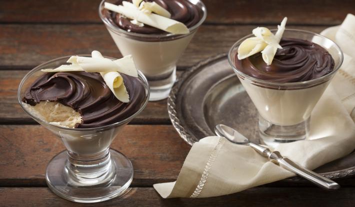 Fotografia em tons de marrom e branco de uma bancada de madeira, sobre ela uma bandeja de metal com um paninho branco e uma taça de vidro com o mousse. Ao lado duas taças de vidro com o mousse.