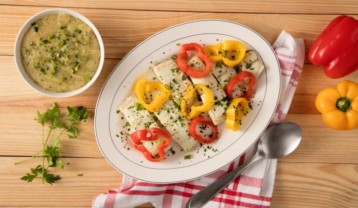 Fotografia em tons de vermelho e amarelo em uma bancada de madeira clara com um pano branco com listras vermelhas. Ao centro, um recipiente branco oval com as postas de peixe e pimentões amarelos e vermelhos dentro dele. Ao fundo, um pote com o pirão.