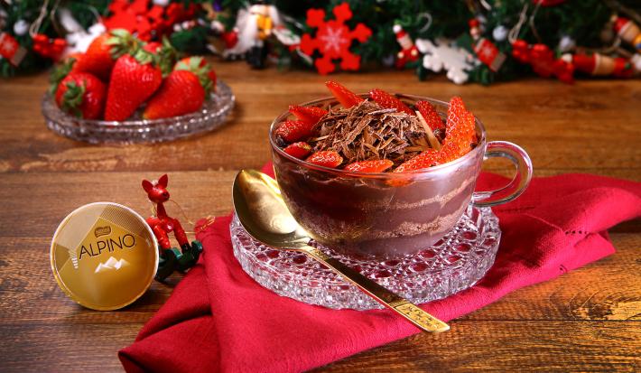 Fotografia em tons de vermelho em uma bancada de madeira, um pano vermelho com uma taça de vidro e o pavê de chocolate Alpino com morangos dentro dela. Ao lado, uma colher dourada e uma cápsula de Dolce Gusto Alpino. Ao fundo, decoração natalina.