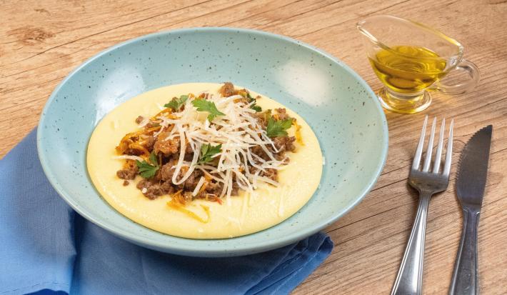 Fotografia de um prato fundo azul claro com uma polenta cremosa, e por cima, uma carne moída com cheiro verde e queijo parmesão ralado. O prato está apoiado em um pano azul pequeno que está sobre uma mesa de madeira, e ao lado tem talheres e azeite.