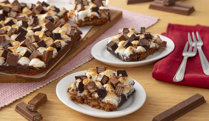 Fotografia em tons de marrom em uma bancada de madeira de cor bege. AO centro, 2 pires contendo os s'mores. Ao fundo, um pano vermelho contendo dois garfos, um pano com bolinhas vermelhas contendo uma tábua mais s'mores e alguns kikat espalhados.