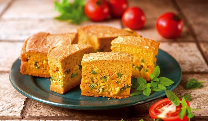 Fotografia de fatias de um bolo salgado em um prato, recheado com vegetais como milho e ervilhas. Tomates frescos e ervas verdes decoram a apresentação.