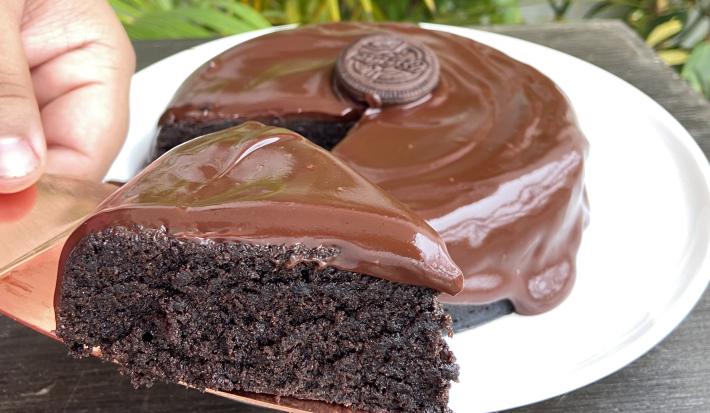 Foto aproximada de um bolo de chocolate com a fatia na frente sobre uma faca. O bolo está decorado com uma calda de chocolate e com um biscoito Negresco no centro.
