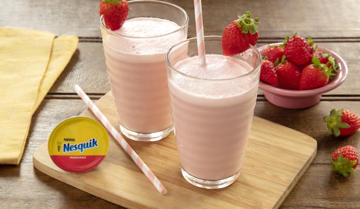 Fotografia em tons de rosa, em uma bancada de madeira de cor marrom. Ao centro, uma tábua de madeira contendo o Frozen. Ao lado, há uma cápsula de Nesquik e um pano amarelo. Ao fundo, há um recipiente com alguns morangos espalhados.