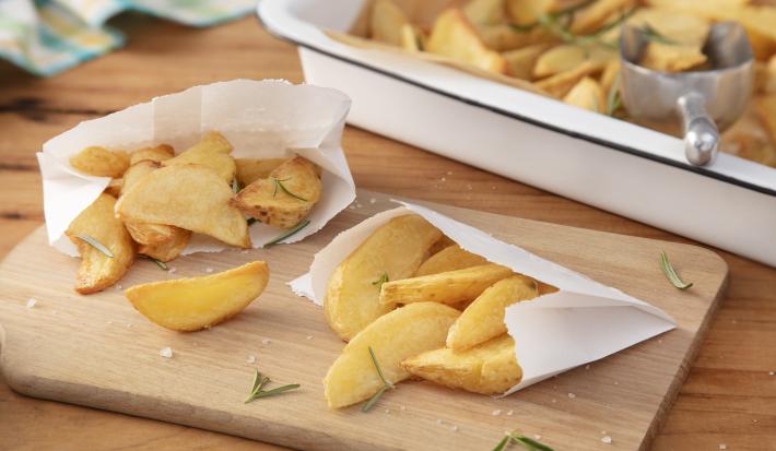 Imagem de dois saquinhos brancos de papel com batatas fritas rústicas dentro sobre uma tábua de madeira. Ao fundo, uma travessa branca com mais batatas e um pegador.
