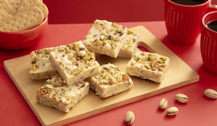 Foto da receita de Fudge de Pistache com TOSTINES Maisena e GALAK. Observa-se uma decoração vermelha com uma tábua de madeira e o fudge cortado por cima. Pistaches decoram a foto.