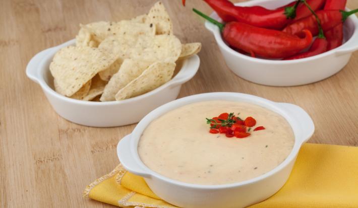 Fotografia em tons de amarelo em uma bancada de madeira clara, um paninho amarelo, um potinho branco com o patê de batata, outro potinho branco com tortillas e ao fundo um potinho com pimenta de dedo-de-moça.