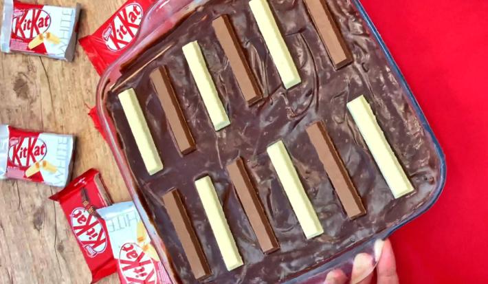 Foto de um pavê de KitKat ao leite e branco, numa travessa de vidro, sobre um tecido vermelho com alguns pacotes de KitKat ao fundo.
