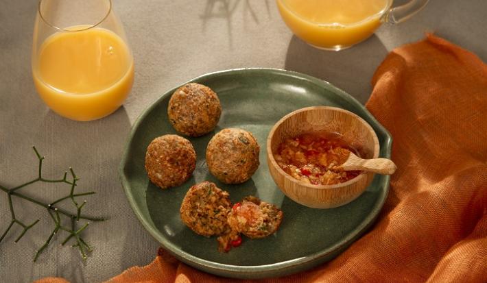 Foto em tons de laranja e verde escuro da receita de bolinho de arroz e aveia servido em 4 porções com uma cumbuca de geleia de pimenta ao lado, tudo sobre um prato de cerâmica verde. Ao lado há um paninho laranja e dois copos de suco de laranja