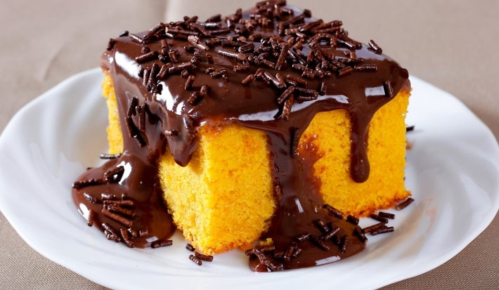 Um pedaço de bolo de cenoura também em formato quadrado, com calda de chocolate escorrendo generosamente pelas laterais e decorado com granulados de chocolate. Ele está servido em um prato branco sobre uma toalha ou superfície marrom.